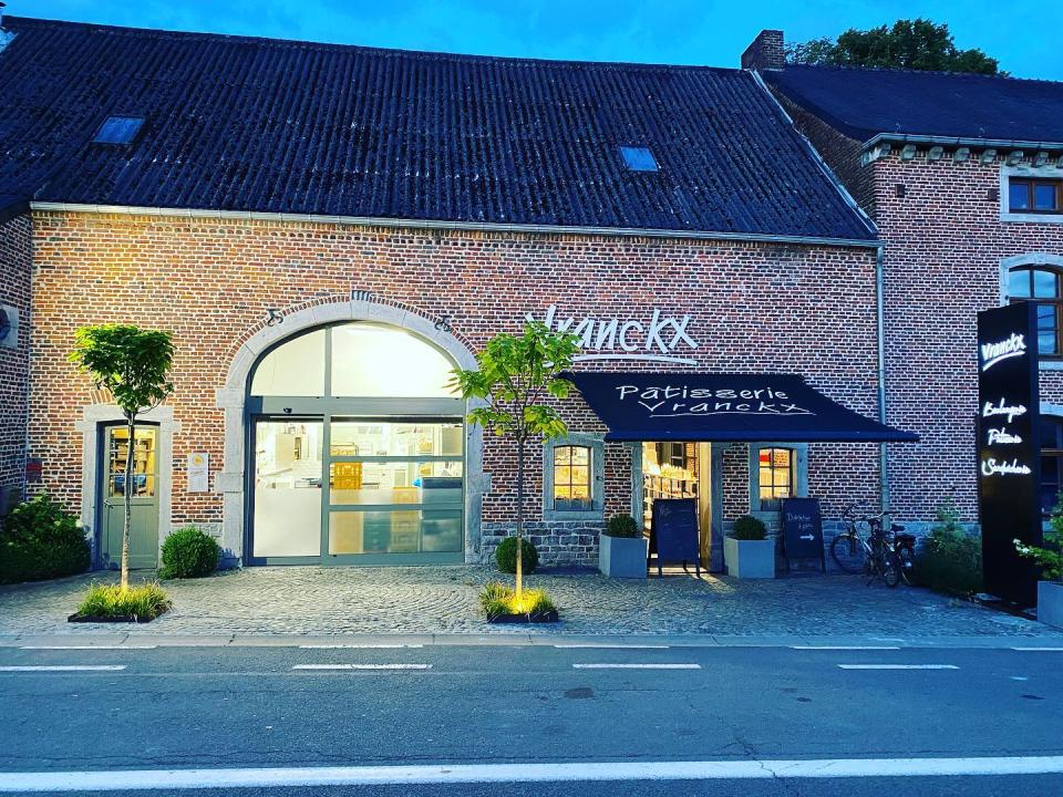 Boulangerie P&#xe2;tisserie Vranckx, a brick building lit from inside, flanked by two trees on cobblestones.