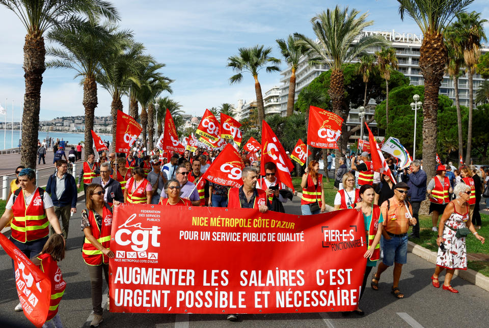 French labour union workers march on a palm-lined street, waving crimson flags and matching banner.