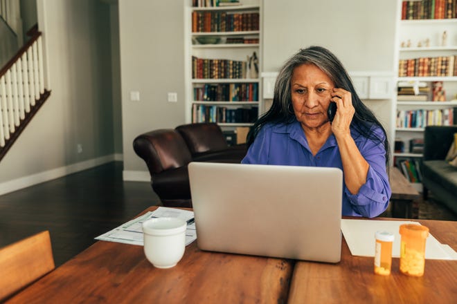 A person looks at a laptop and uses a phone while sitting at a table at home.