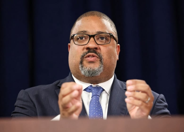 Close up of individual with short dark hair wearing glasses, speaking at a podium, gesturing with his hands.