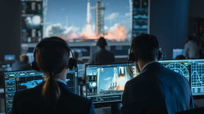 A group of people wearing headsets in front of desktops inside a control center witness a rocket ship launch.