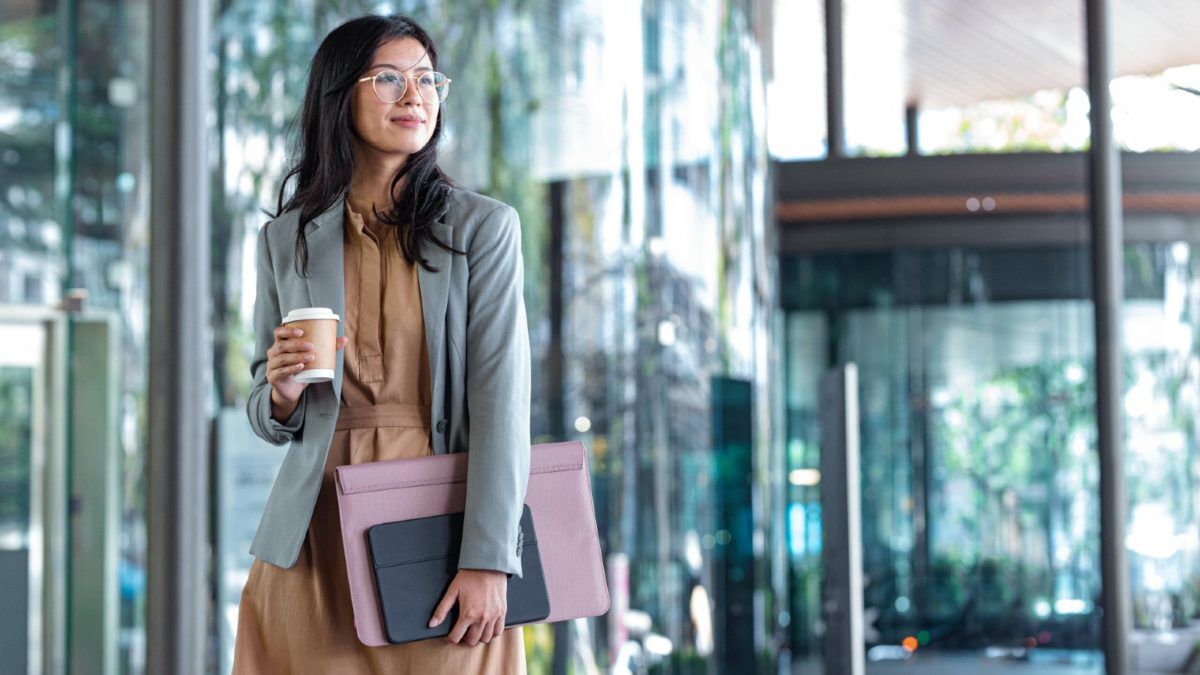 Young Asian woman holding a cup of takeaway coffee and folders containing paperwork, on her way into the office