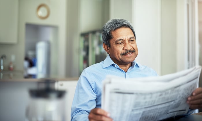A smiling person reading a financial newspaper while seated at a table in their home.