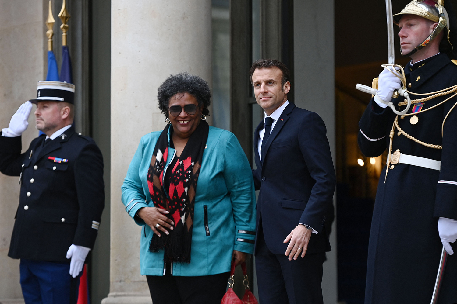 French President Emmanuel Macron, wearing a dark suit and tie, welcomes Mia Mottley, the prime minister of Barbados, wearing a teal jacket and colorful scarf, before a meeting in Paris. They are flanked by French guards in uniform.