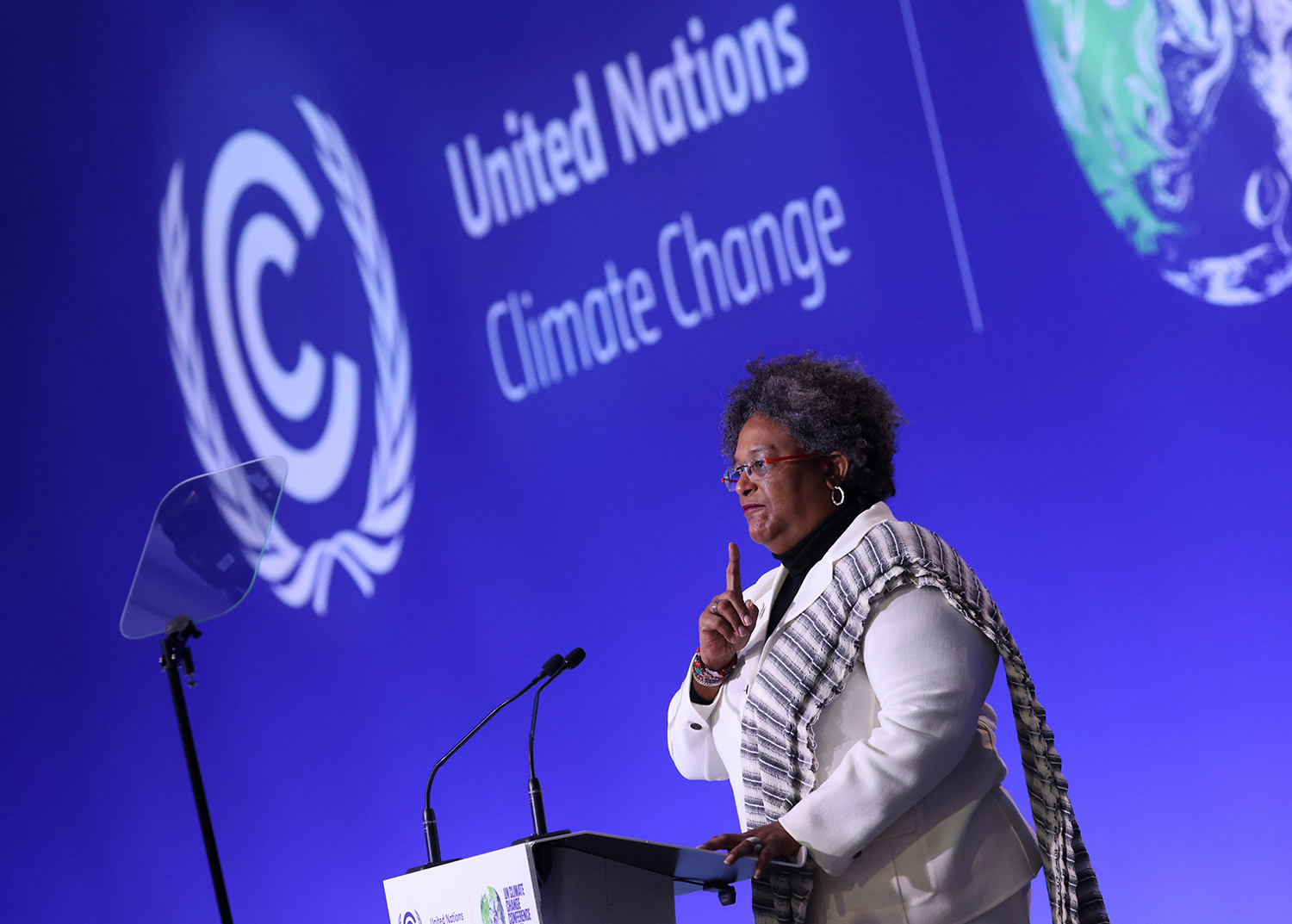 Mia Mottley, the prime minister of Barbados, wearing a white suit with patterned fabric on the shoulder, speaks from a podium under a sign that reads "United Nations Climate Change."