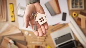 Image of a man holding a key chain with a key and house attached to the key ring over a office desk in the background