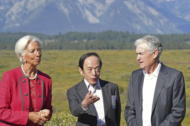 Woman and two men in suits stand in a line talking in front on green grass, trees and large mountains in the background.