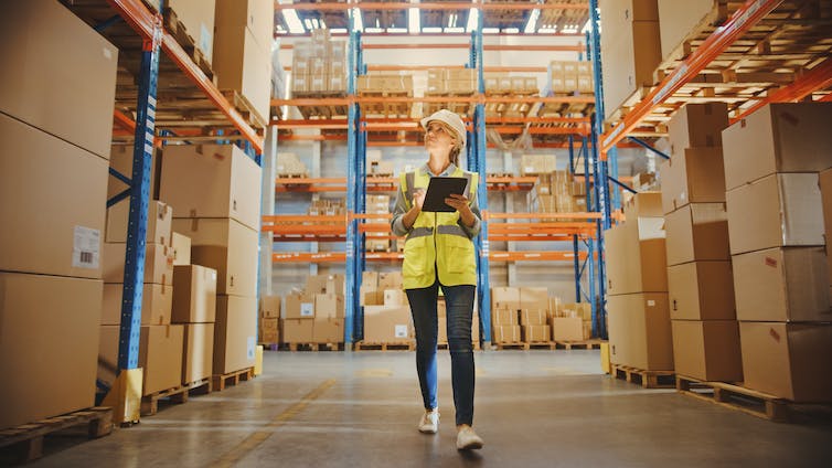 Woman in high-vis vest and hard hat walks through a warehouse full of boxes with a tablet.