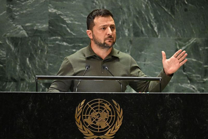 Ukrainian President Volodymyr Zelensky, wearing a dark green shirt, gestures with his left hand, as he stands behind the U.N. seal to address the United Nations General Assembly in New York.