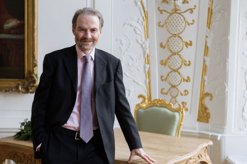 Timothy Garton Ash, wearing a suit and tie, rests his hand on a wooden table. Behind him is a gilded chair and ornate wall with a large gilt frame partially seen at left.