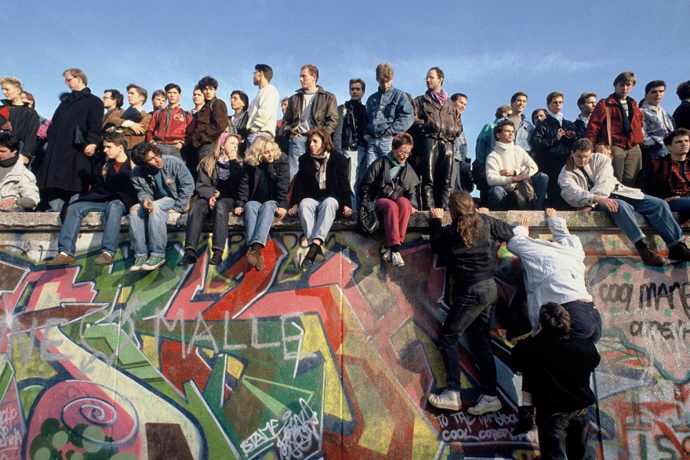 A crowd made up mostly of young people sit, stand, and climb the graffiti-covered Berlin Wall in 1989.