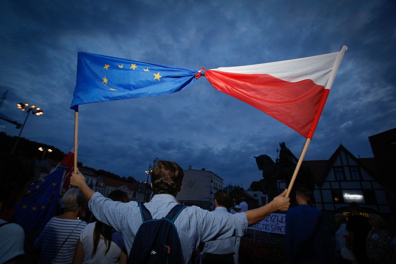 A main in a striped collared shirt, wearing a backpack, holds an EU flag intertwined with a Polish flag during a solidarity rally in Bydgoszcz, Poland. He is seen against a darkening, cloudy twilight sky with a crowd of others and a statue in the distance.