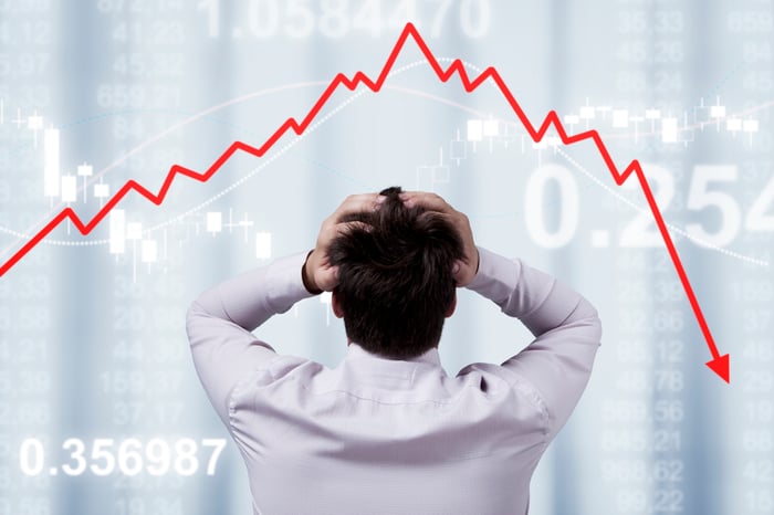 A person gripping their head in frustration while watching a stock market trend line move lower.
