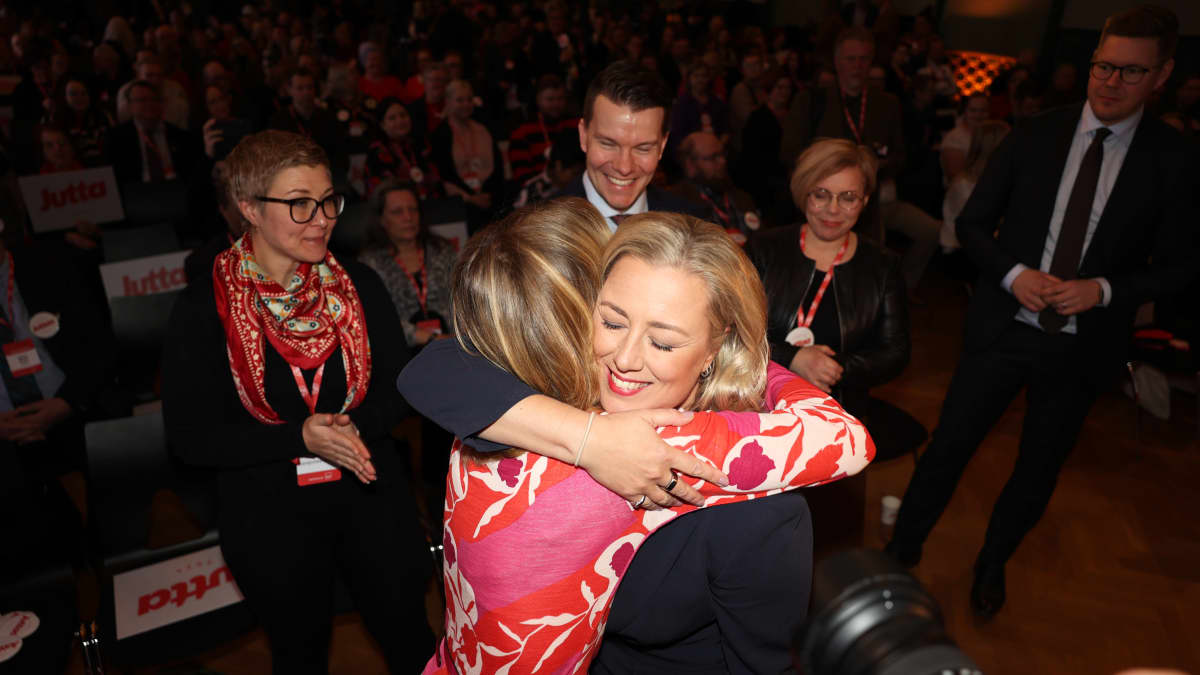 Two women hugging as four other smiling, formally dressed people stand behind them and others are seated in a dark auditorium.