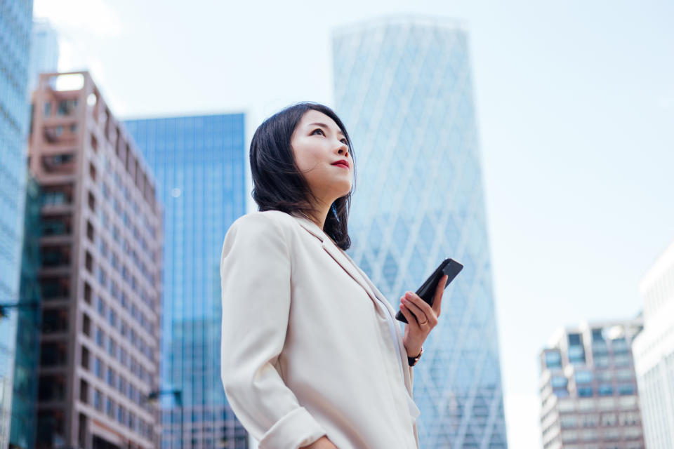 women Low angle view of beautiful young Asian woman using smartphone while standing in front of commercial buildings in financial district. Young professional looking for a new career opportunity.