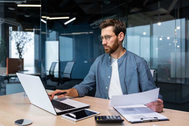 Serious and focused financier accountant on paper work inside office, mature man using calculator and laptop for calculating reports and summarizing accounts, businessman at work in casual clothes