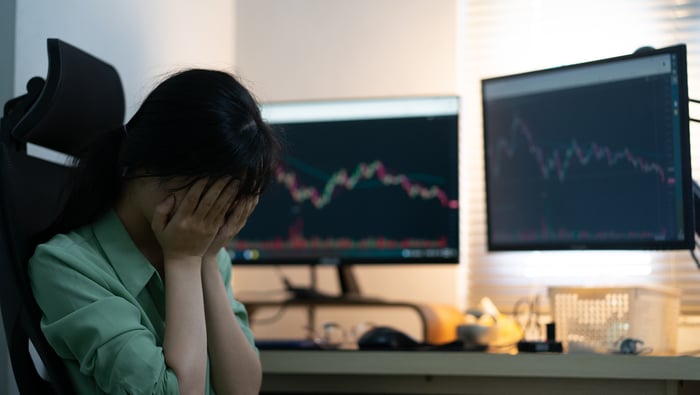 A person with hands over face sitting next to screens showing declining stock charts.