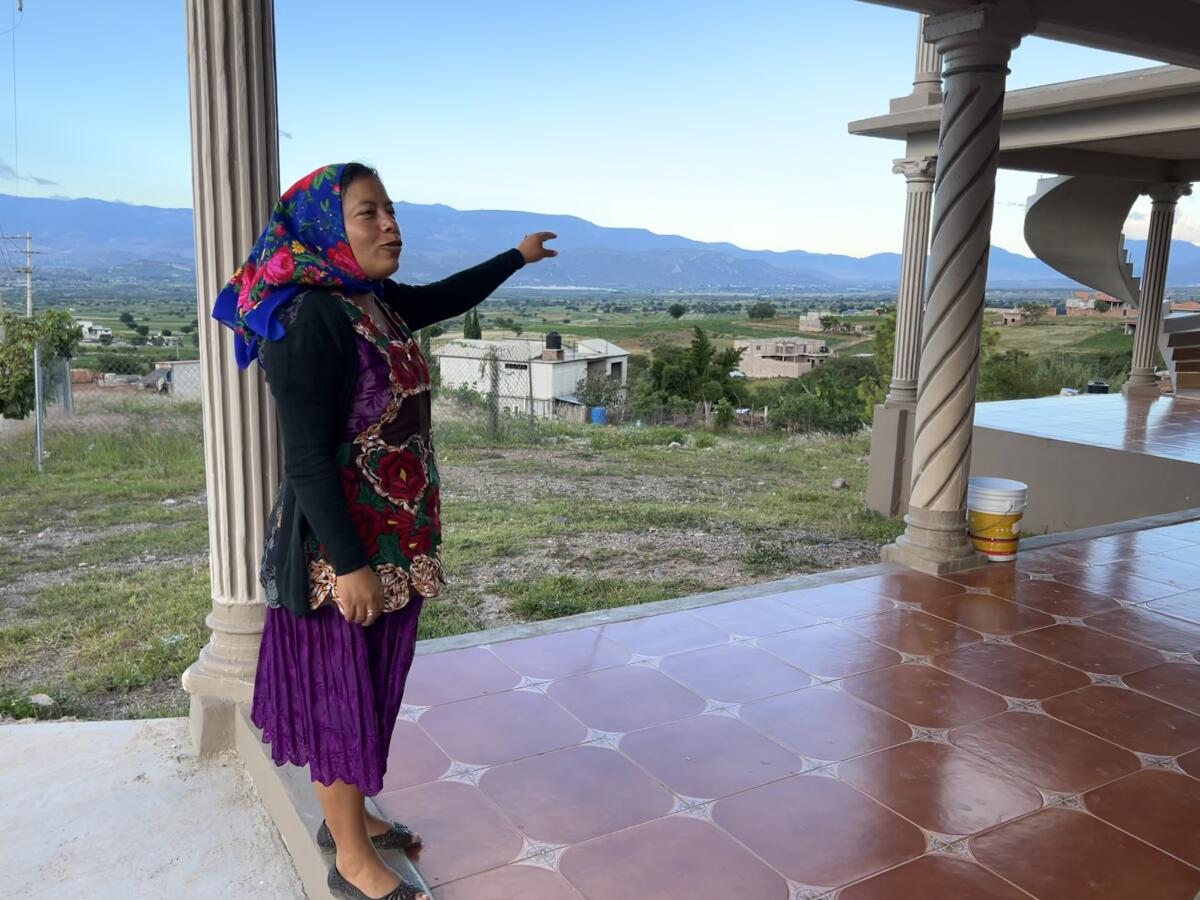 A woman stands in a tiled, outdoor room with columns, with mountains in the background.