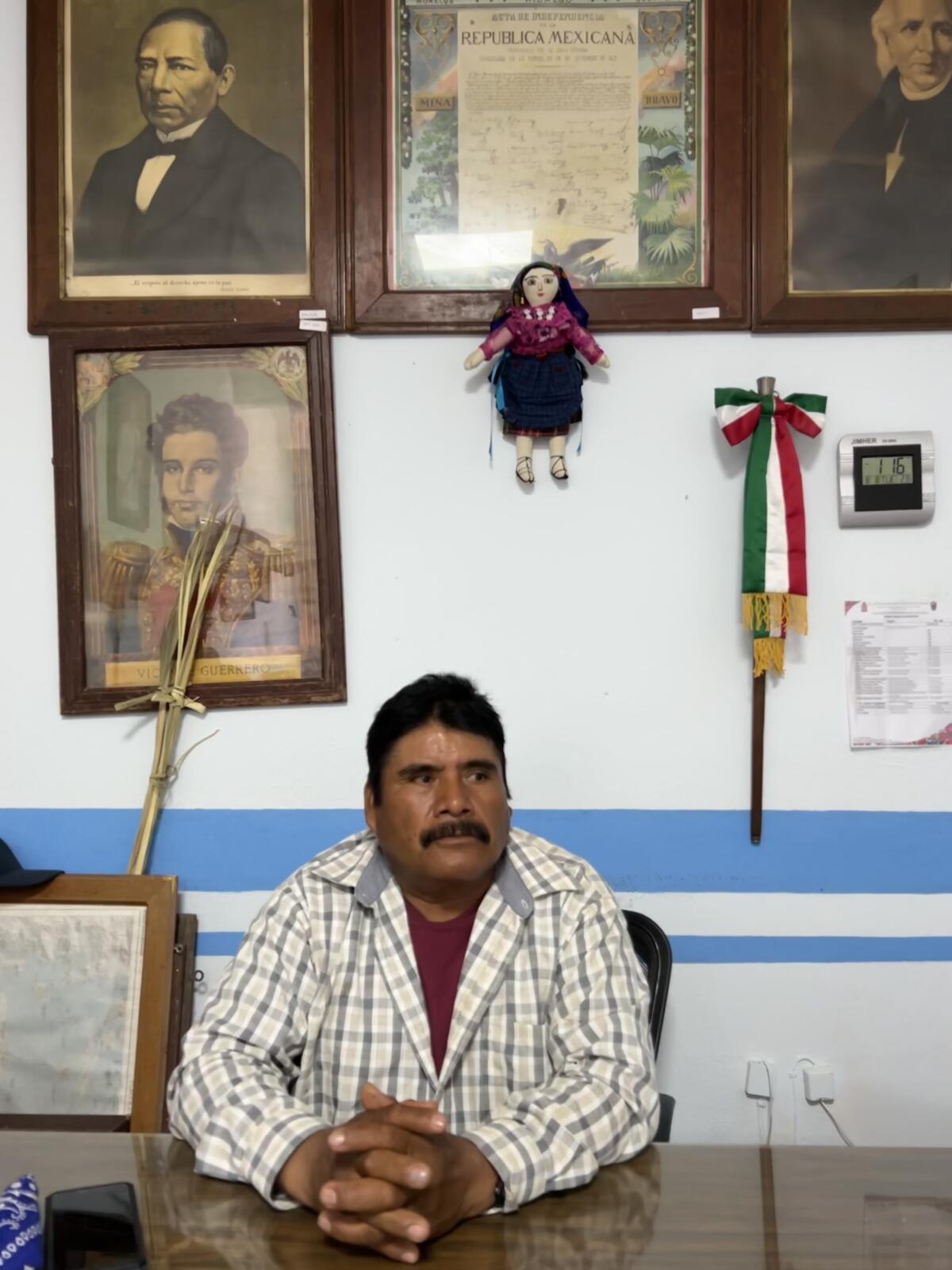A man sits at a desk in front of a wall with historic Mexican portraits hanging on it. 