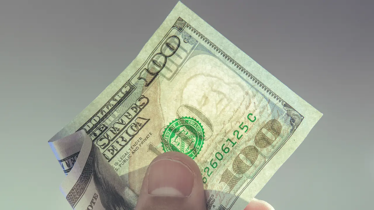 A hand holding a one hundred dollar bill with translucent Franklin watermark visible.