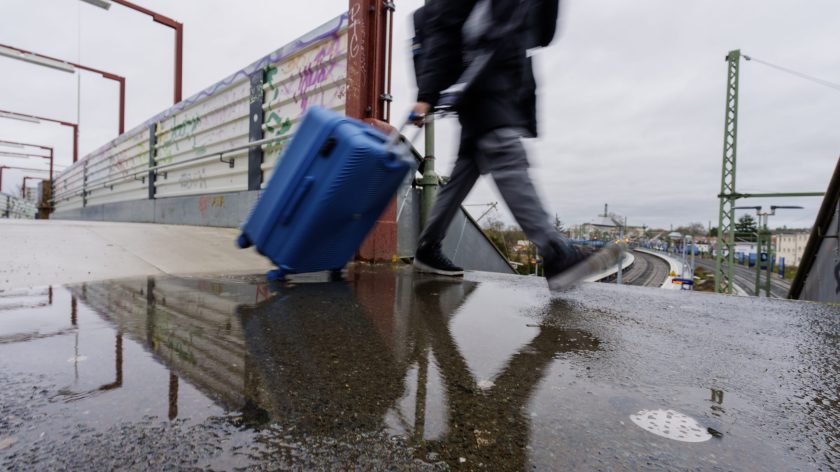 A blurred image of a person whose face is not visible, lugging a blue cabin suitcase.