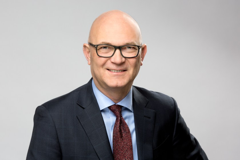 A bald man wearing glasses and a suit, smiles and looks ahead.