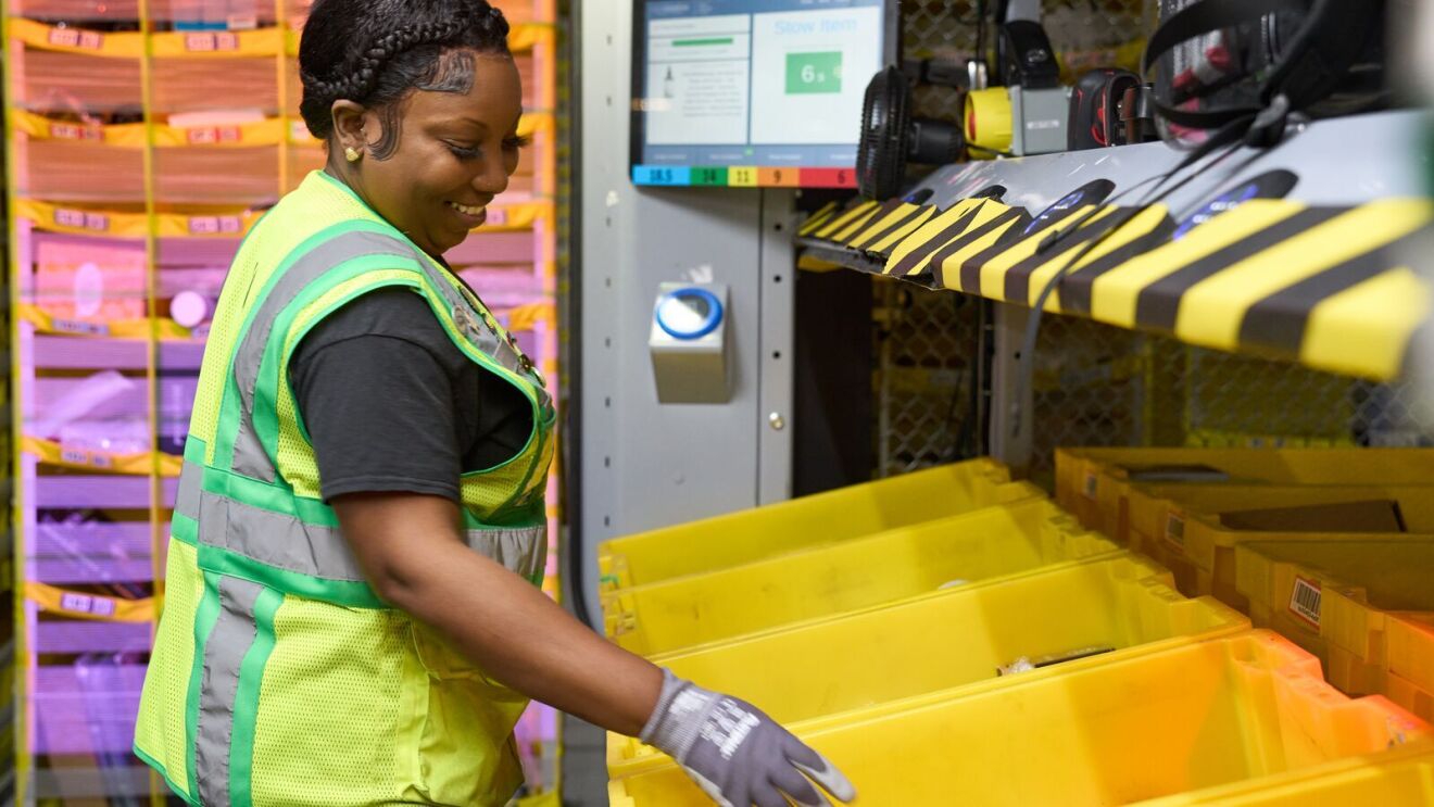 A photo of an Amazon employee who works at a fulfillment center checking products in shipment bins on a shelf.