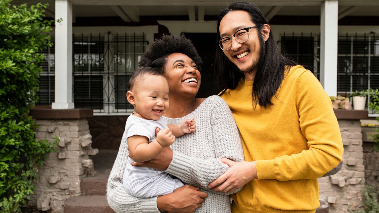 A couple smiles and poses with their baby in front of their home.