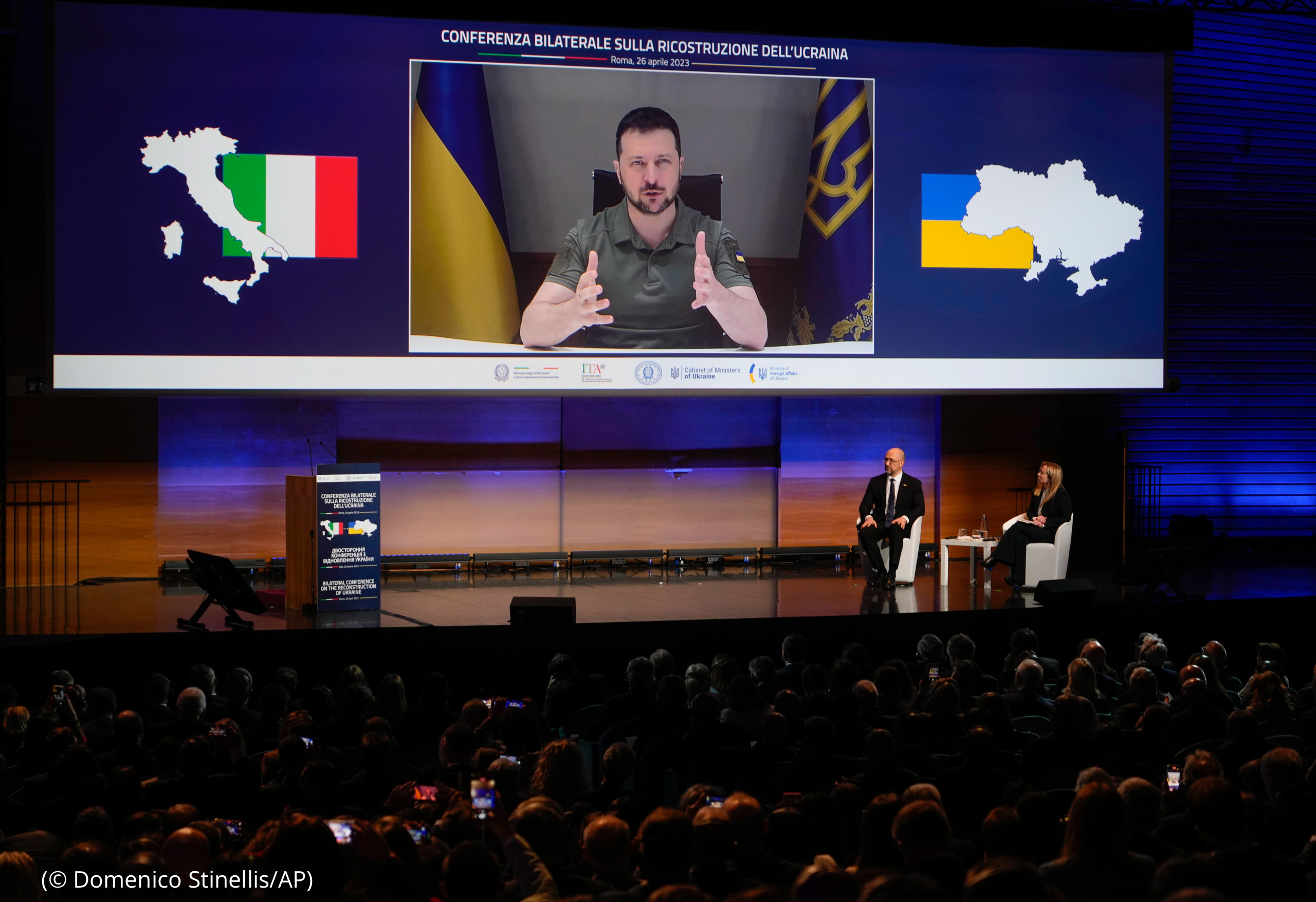 Volodymyr Zelenskyy speaking on large screen above stage in auditorium (© Domenico Stinellis/AP)