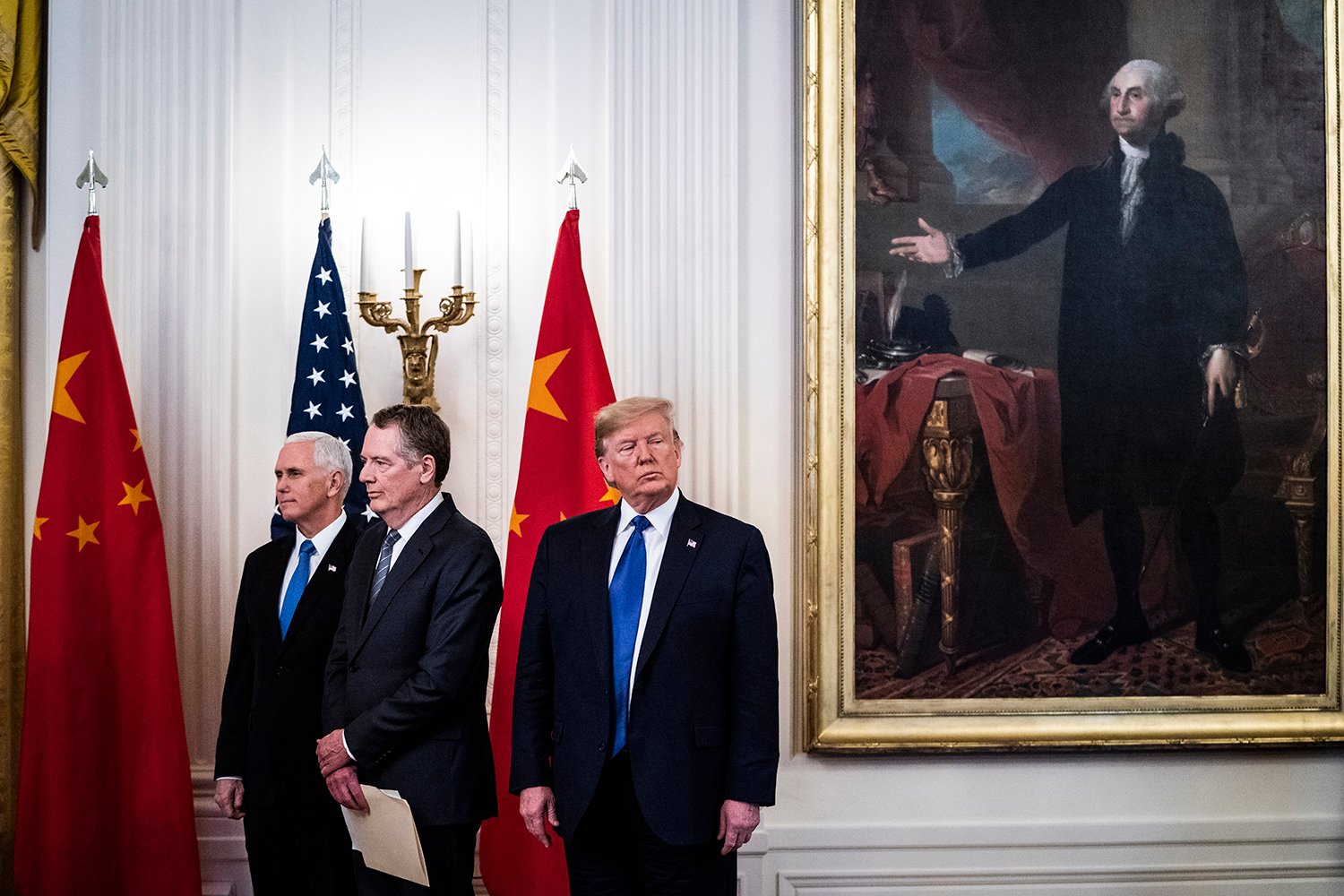 Three men in suits and ties stand in front of two Chinese and one U.S. flag. Behind them is an ornate wall and larger portrait of George Washington.