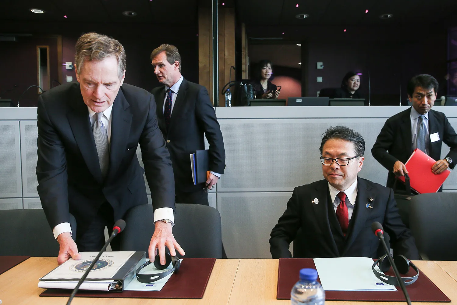 Robert Lighthizer, (left) in a suit and tie, leans over to place a binder and headsphones on a table in front of him as he prepares to sit next to the seated Japanese economy minister, also wearing a suit and tie. Behind them are two other staffers. Two other women are seen seated behind a low wall in the background.