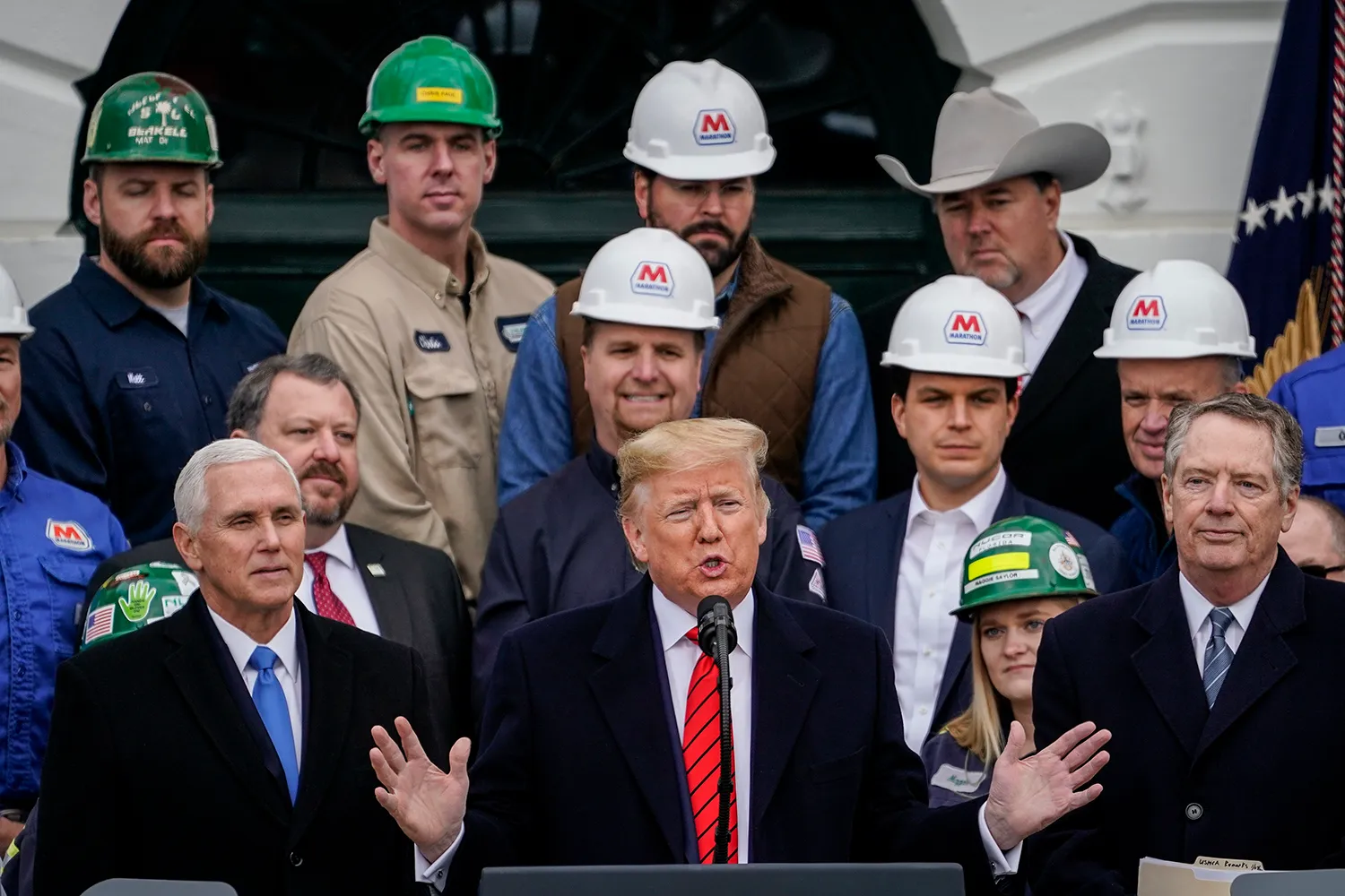 Men and a woman in hardhats and one man in a cowboy hat stand behind Pence, Trump, and Lighthizer, all in suits and ties. Trump gestures with both hands as he speaks in front of a microphone.