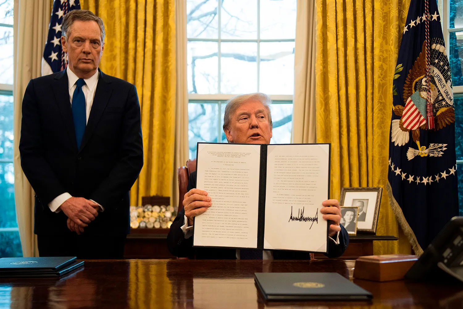 Lighthizer in a suit and tie stands behind a seated Trump who holds up a two-page document with his signature on it. Behind them are curtains, window and flag of the Oval Office.