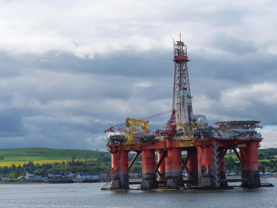 An oil drilling platform stands in the calm sea, behind it a cloudy sky and green landscape, drilling platform in red against a blue sky in the north