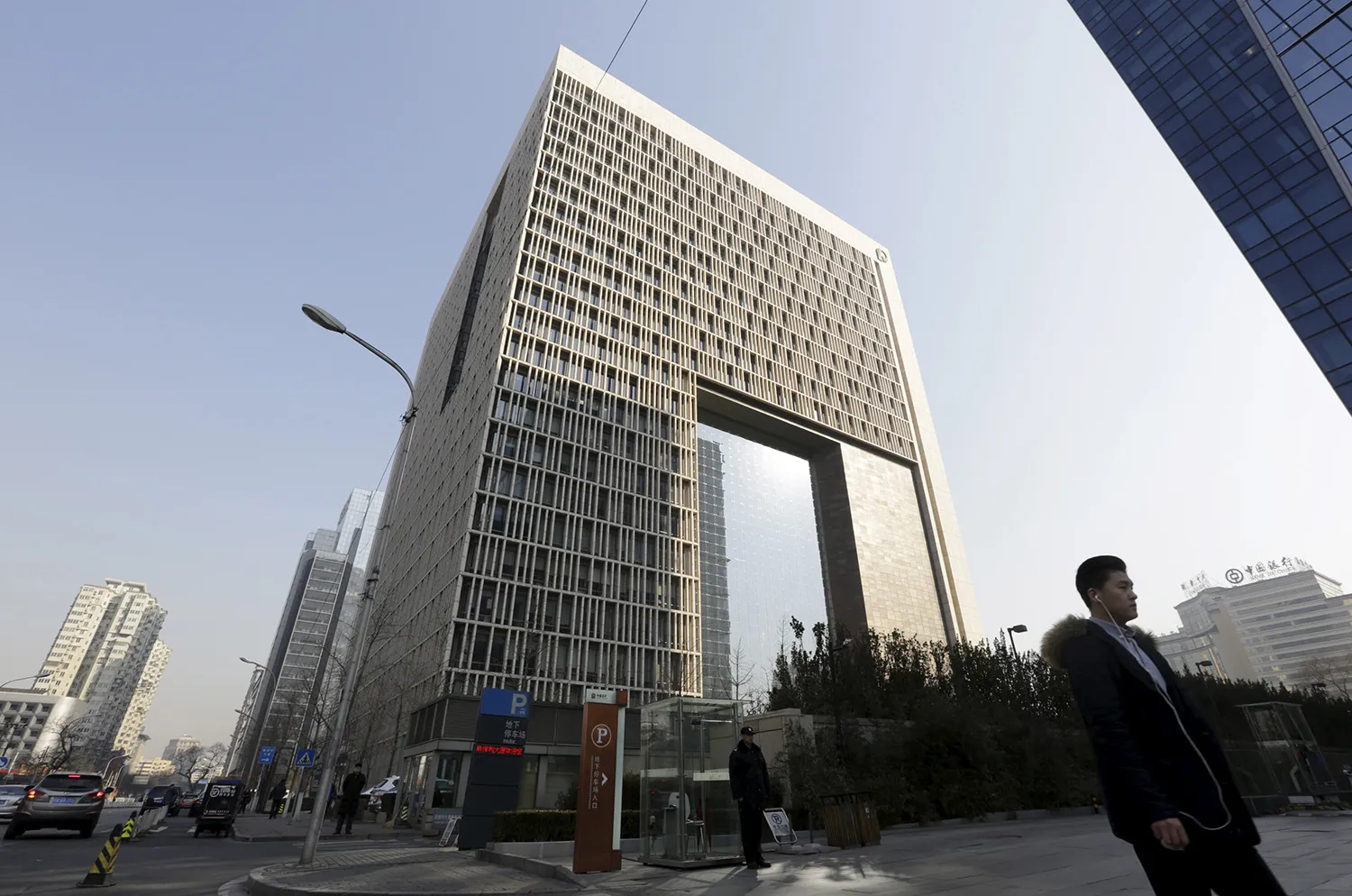 A man wearing headphones walks on a street past a tall building.