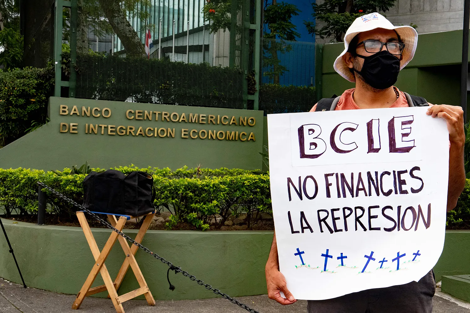 A Nicaraguan citizen exiled in Costa Rica, who wears a bucket hat and a protective face mask, holds a sign that reads: "BCIE: No Financies La Represion" in front of the CABEI bank building during a protest.