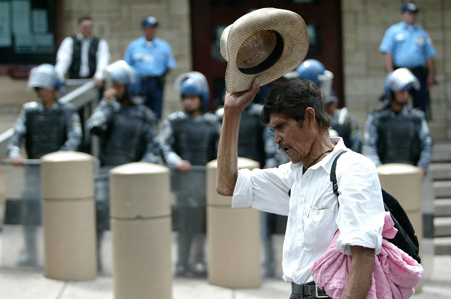A Honduran man wearing a white shirt with sleeves rolled up and carrying a backpack lifts his straw hat and shouts as he protests in front of the U.S. Embassy in Tegucigalpa. Behind him and out of focus are traffic barriers and riot police in full gear and helmets.