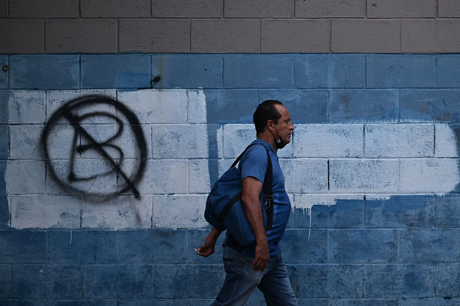 A man in a T-shirt with a face mask around his chin and carrying a backpack walks past a cinderblock wall that has been spray-painted with an anti-bitcoin symbol showing a crossed out letter "B."