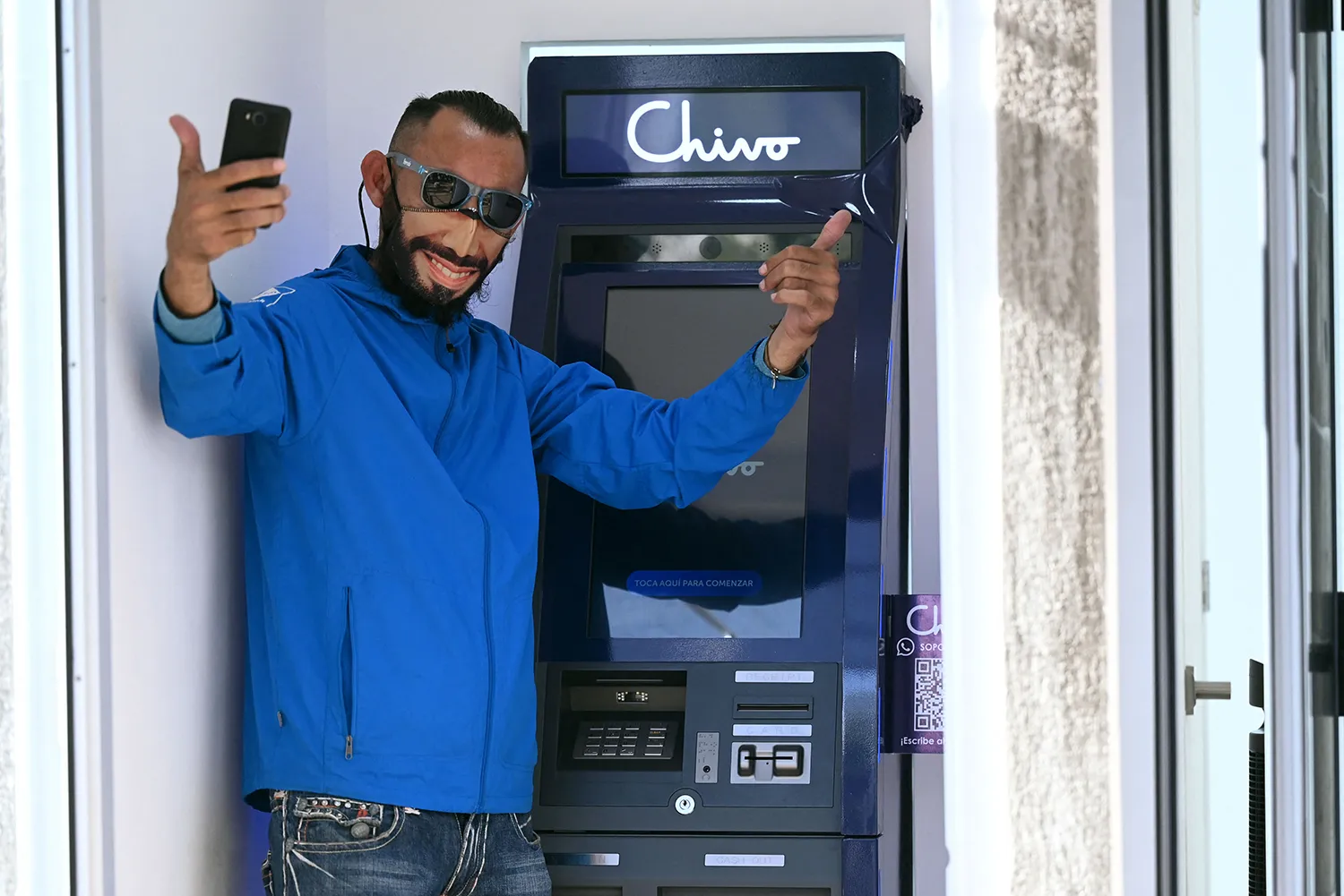A man wearing sunglasses and a cloth face mask with the image of Salvadoran President Nayib Bukele's face on it poses with two thumbs up as he stands in front of a Chivo bitcoin ATM.