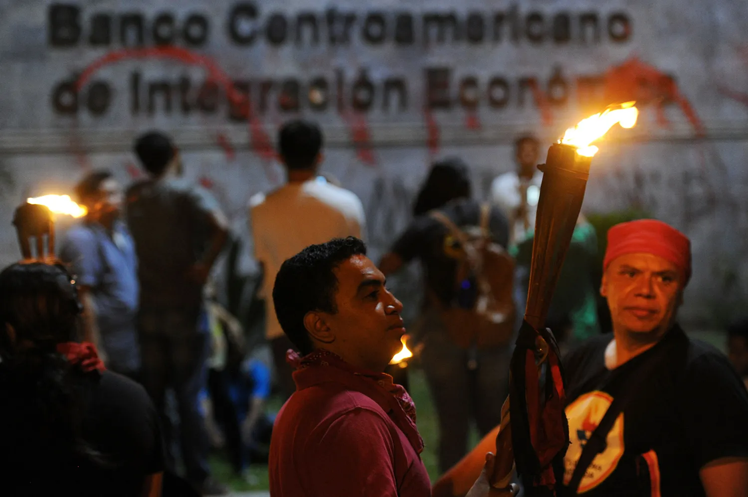 A man holding a lit torch stands among a small crowd of protesters outside the Central American Financial Integration Bank in Tegucigalpa. A red paint-splattered sign for the bank is seen behind the demonstrators.