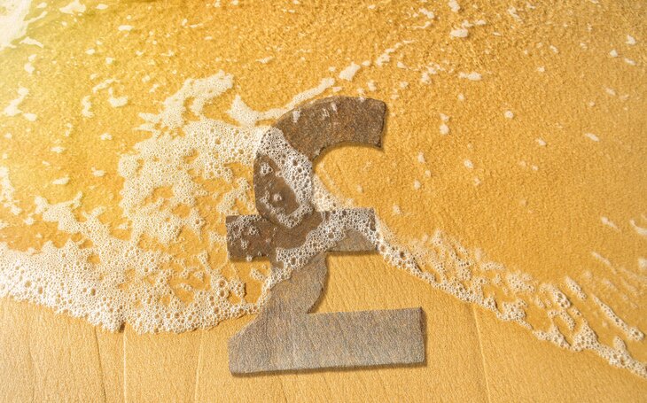 A stone UK pound sign lies on a sandy beach being washed by salty water in AML.