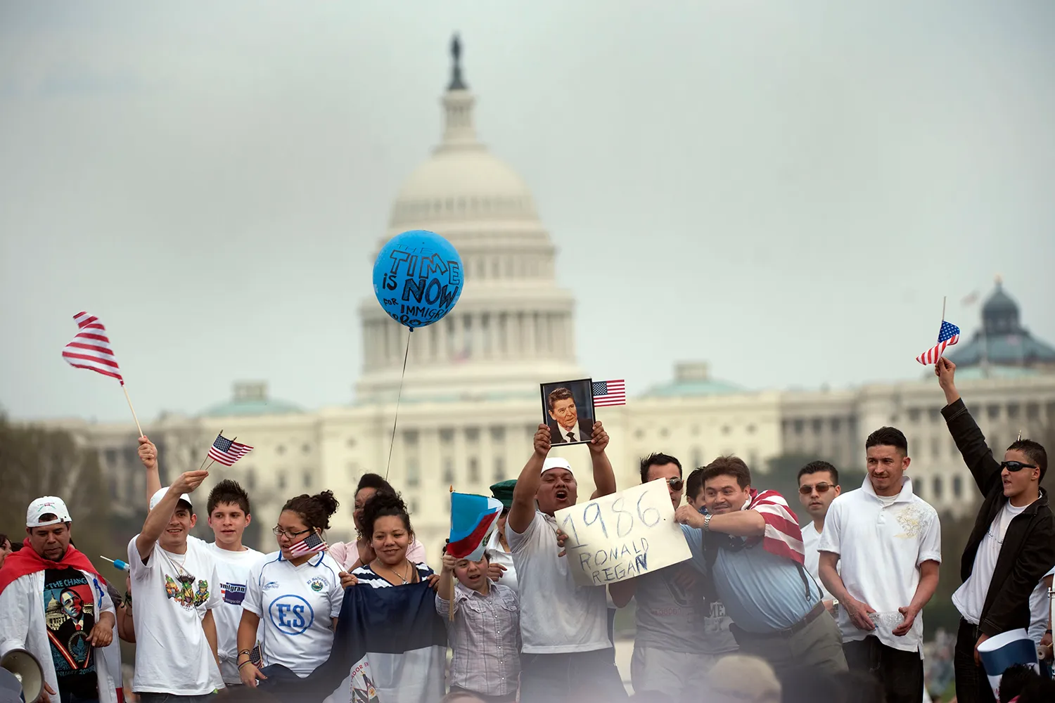 About a dozen people wave flags and signs as they attend a rally in support of immigration rights on the National Mall in Washington, D.C. The white dome of the U.S. Capitol building looms behind them against a pale cloudy sky.