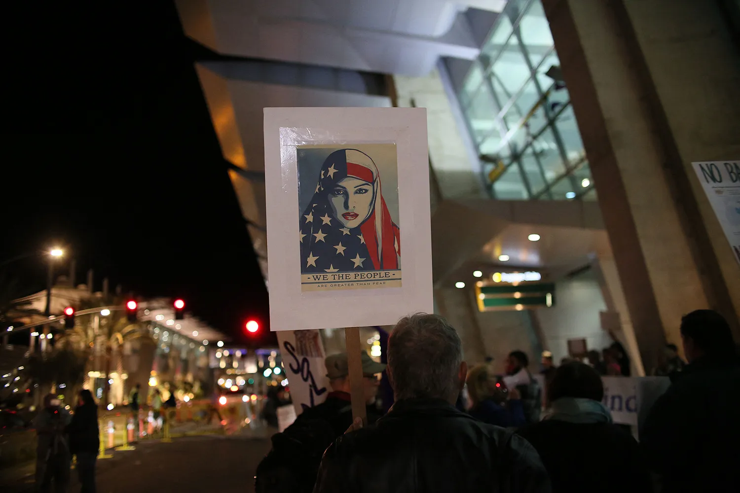 A protester is seen from behind on a sidewalk outside the San Diego airport during a nighttime demonstration. The protester holds a picket sign taped with the image of a woman wearing an American flag as a head scarf. Beneath her is the text: "We the people are greater than fear."