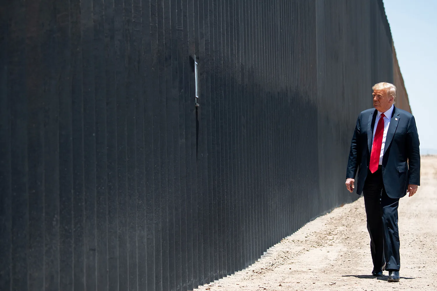 Then-President Donald Trump walks on the dusty ground alongside a long stretch of the U.S.-Mexico border wall, which is black and corrugated and more than twice his height. Trump wears a dark suit and a bright red tie.