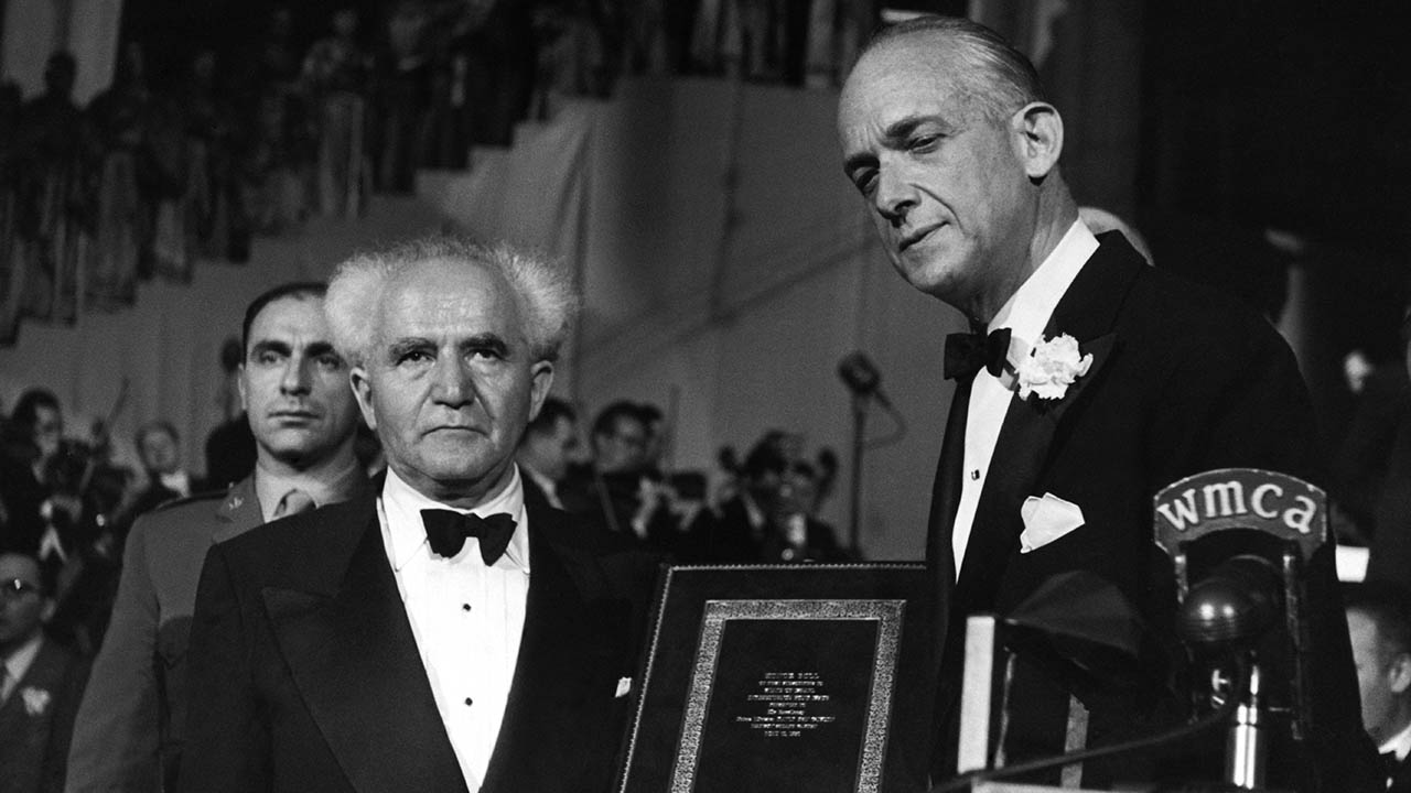 Historic black and white photo showing two men in formal suits holding a small plaque.